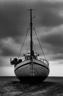 A fishing boat sits dry on a sandy beach in Denmark under dark, dramatic clouds, creating a moody and high-contrast coastal scene.