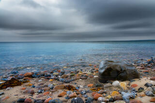 A rocky Danish beach with a large boulder on the shore, while a sailboat drifts under dark, stormy clouds on the horizon.