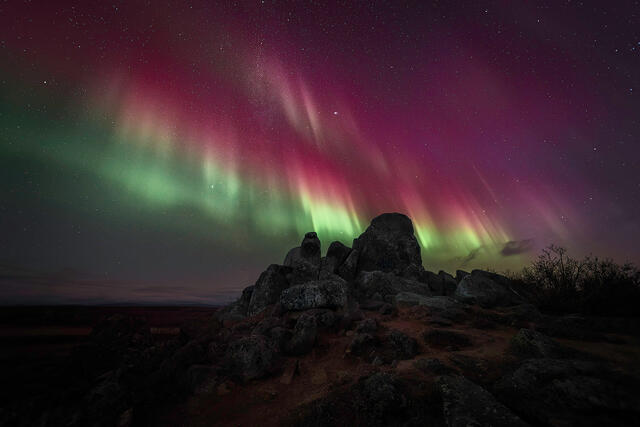 Red and green aurora borealis with the Milky Way arcing over a granite tor outcrop on open tundra in Alaska on a clear autumn night.