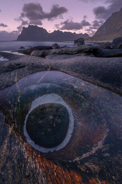 amous Dragon's Eye natural hole formation in rock with tidal pool reflection at Uttakleiv Beach beneath dramatic peaks at moody twilight, Lofoten Islands Norway