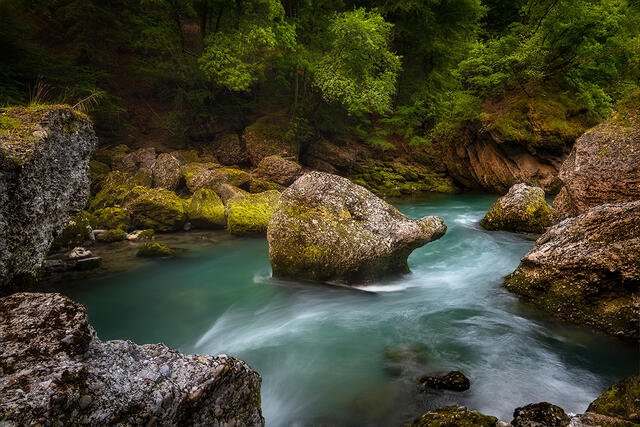 Turquoise glacial water with silky long exposure flowing around massive moss-covered boulder in dramatic rock formations beneath lush green forest, Dranse River