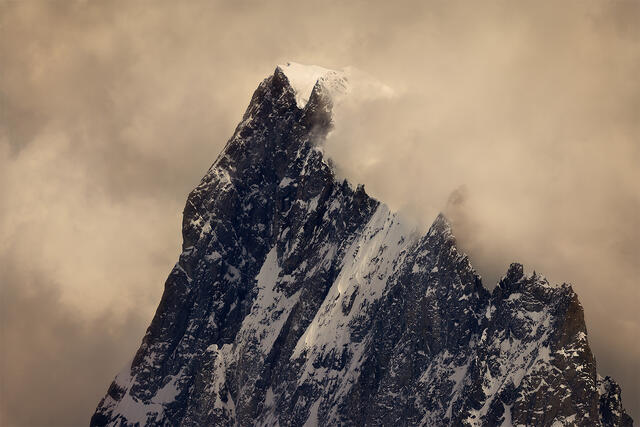 Dramatic jagged Alpine mountain peak with snow and rock emerging from swirling clouds with warm golden atmospheric light in the French Alps France