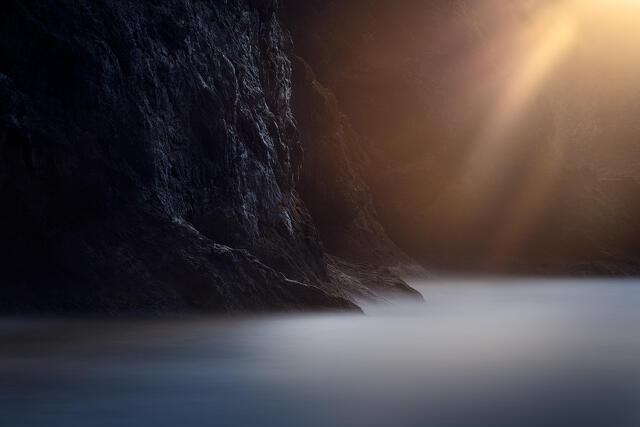 Golden sunbeam over dark rocky coastline with long exposure water, Oregon Coast
