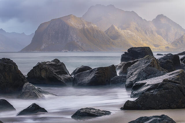 Long exposure of silky water flowing through dark volcanic rocks with dramatic mountain peaks catching golden light in Norway's Lofoten Islands