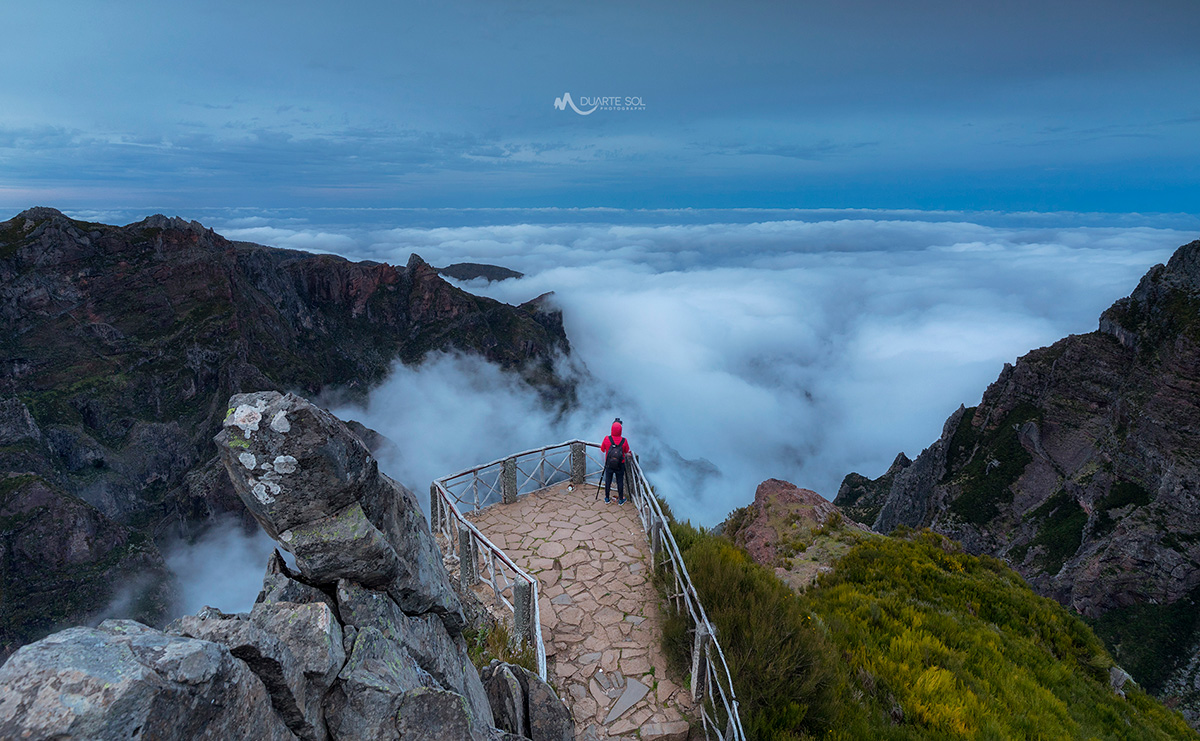 A person standing on a rocky viewpoint with a railing, surrounded by clouds and mountains.