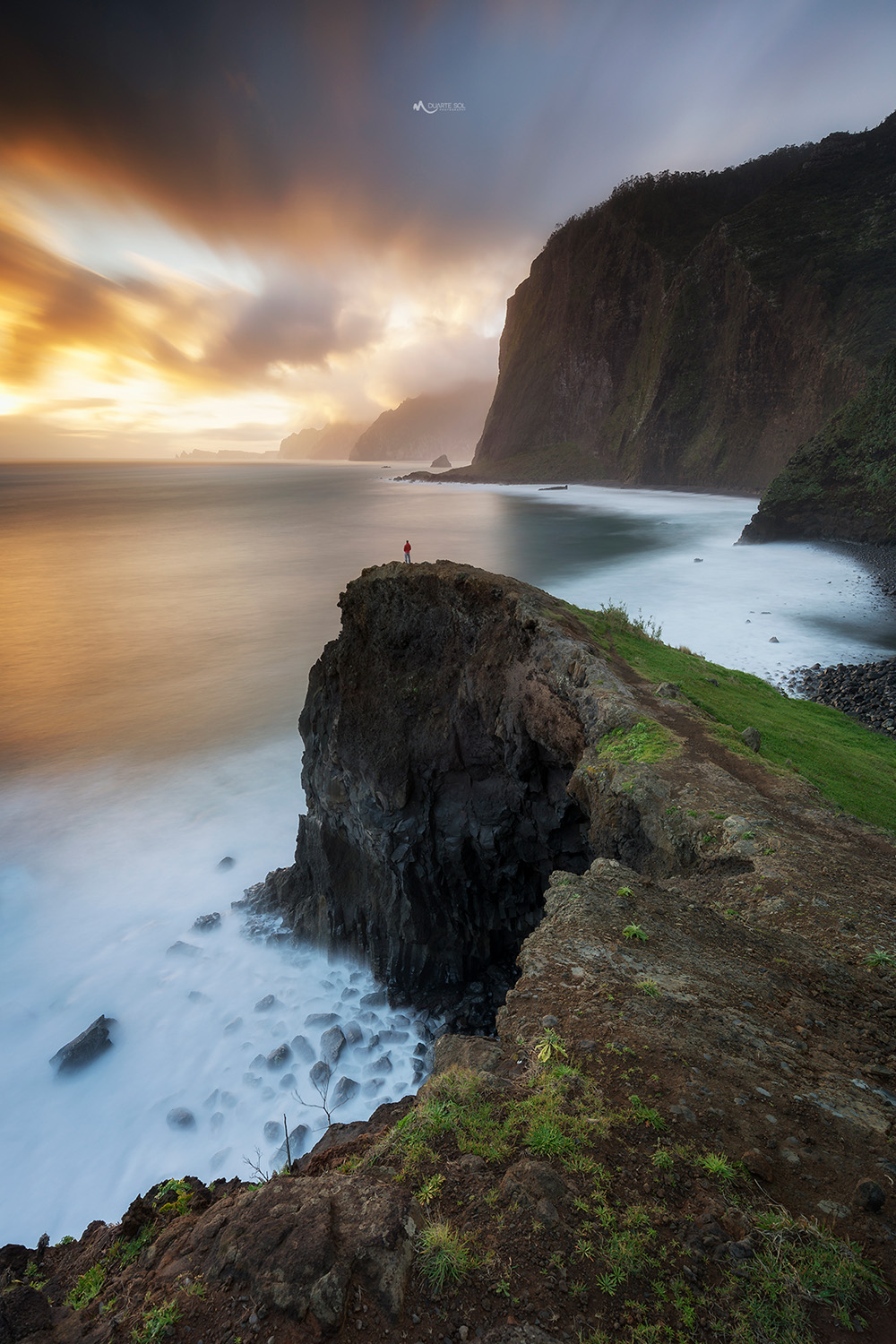 A lone person in red stands on a rocky cliff overlooking a misty ocean at sunset.