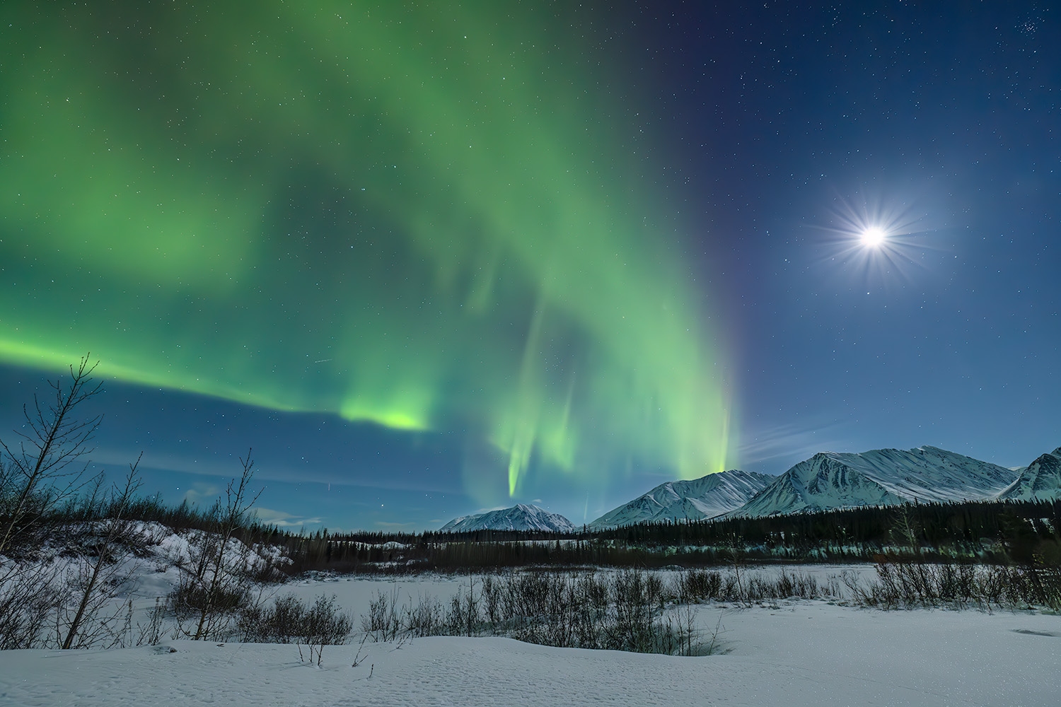 Bright green aurora lights dance above snow-covered mountains and a river under a starry night sky with the moon.
