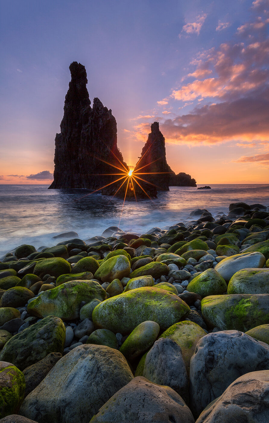 Tall rock formations silhouetted against a colorful sunset, with sunlight shining through. Pebbles and green moss in the foreground.