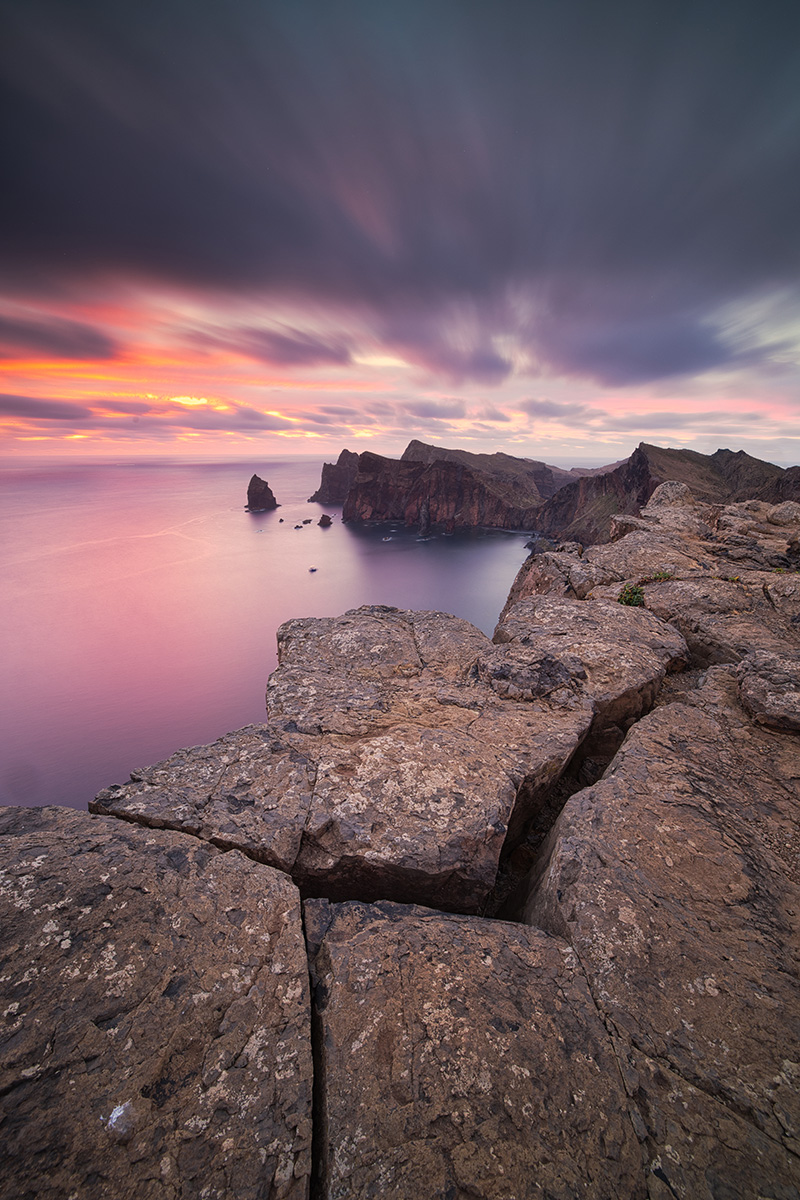 Rugged rocks at a cliff edge overlooking calm waters and distant peaks during sunset.
