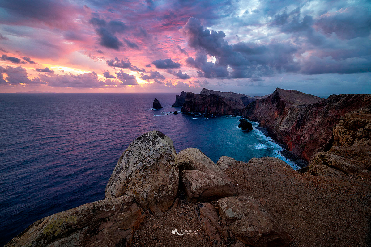 A rocky coastline with cliffs, ocean waves, and a colorful sunset sky.