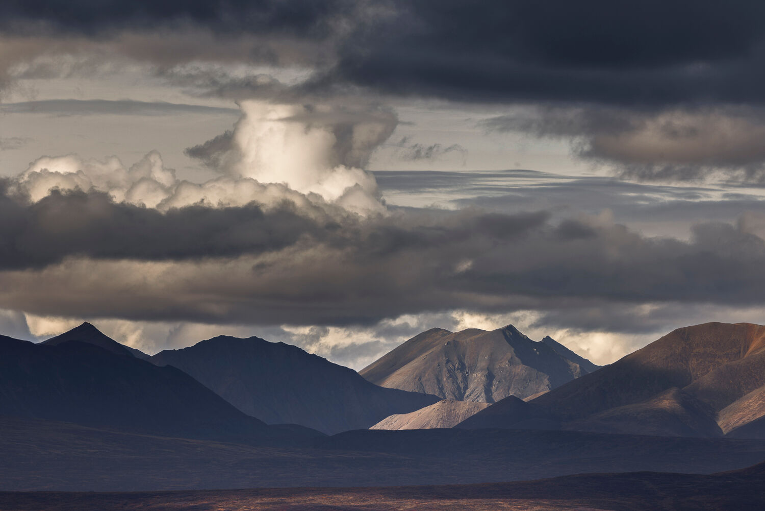 Panoramic Alaskan landscape featuring dramatic cloud formations and rugged mountain peaks during a fall photography workshop. The scene highlights contrasting light and shadow, with illuminated slopes revealing earthy tones and a tundra-like foreground, capturing the raw beauty of Alaska's wilderness.