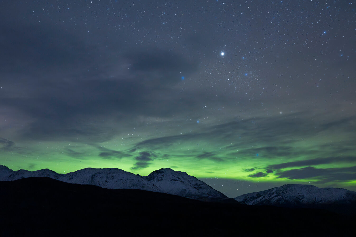 Nighttime view of the Northern Lights over an Alaskan snow-capped mountain range during a fall photography workshop. Vibrant green aurora borealis swirls across a star-filled sky, with rugged peaks silhouetted against the celestial display, highlighting Alaska's natural beauty