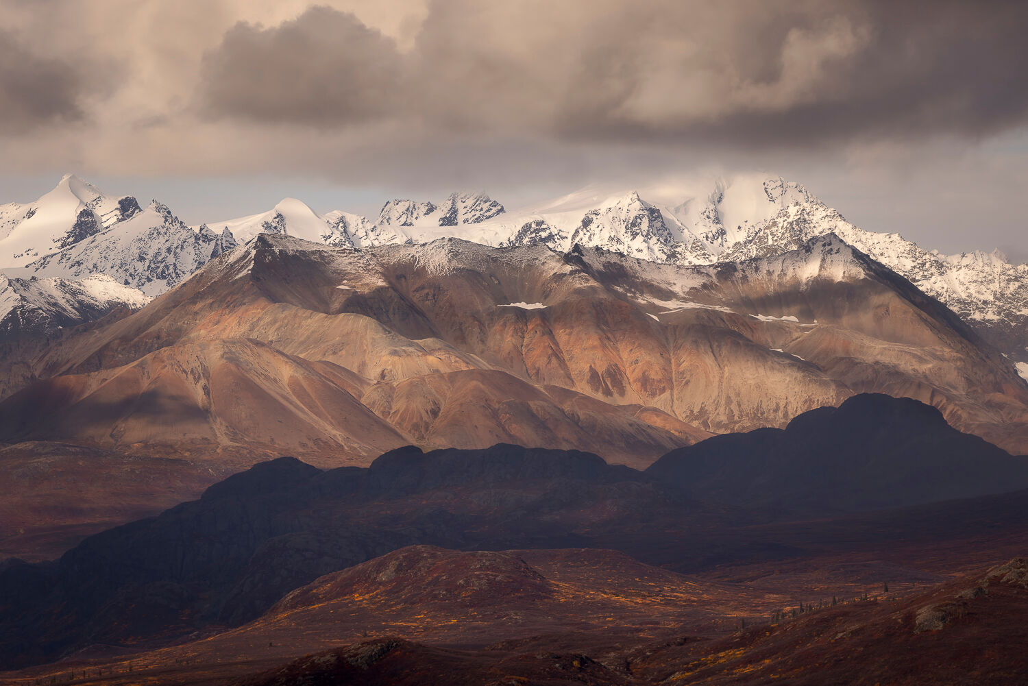Panoramic view of Alaska's rugged autumn landscape during a photography workshop. Snow-capped peaks rise under a dramatic cloudy sky, with terracotta-colored mountains and shadowed, forested hills in the foreground. Sunlight breaks through, highlighting the vibrant fall hues and creating a moody, majestic atmosphere.