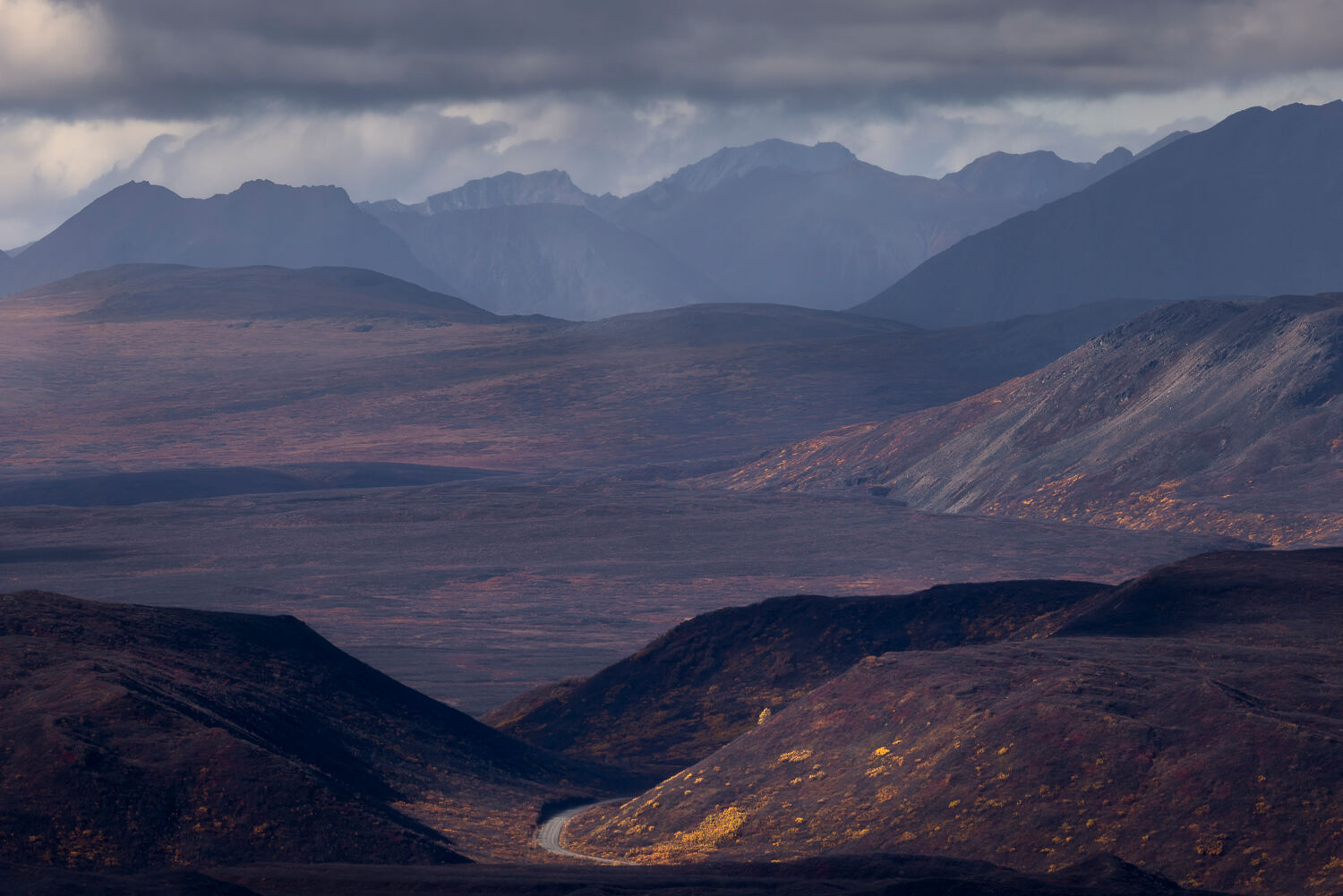 Expansive autumn landscape in Alaska captured during a photography workshop. Layers of rugged mountains and hills stretch under a dramatic cloudy sky, with golden-yellow foliage highlighting the valleys. A winding road in the foreground, illuminated by sunlight, adds depth and scale to the scene, showcasing the region's remote and majestic beauty.