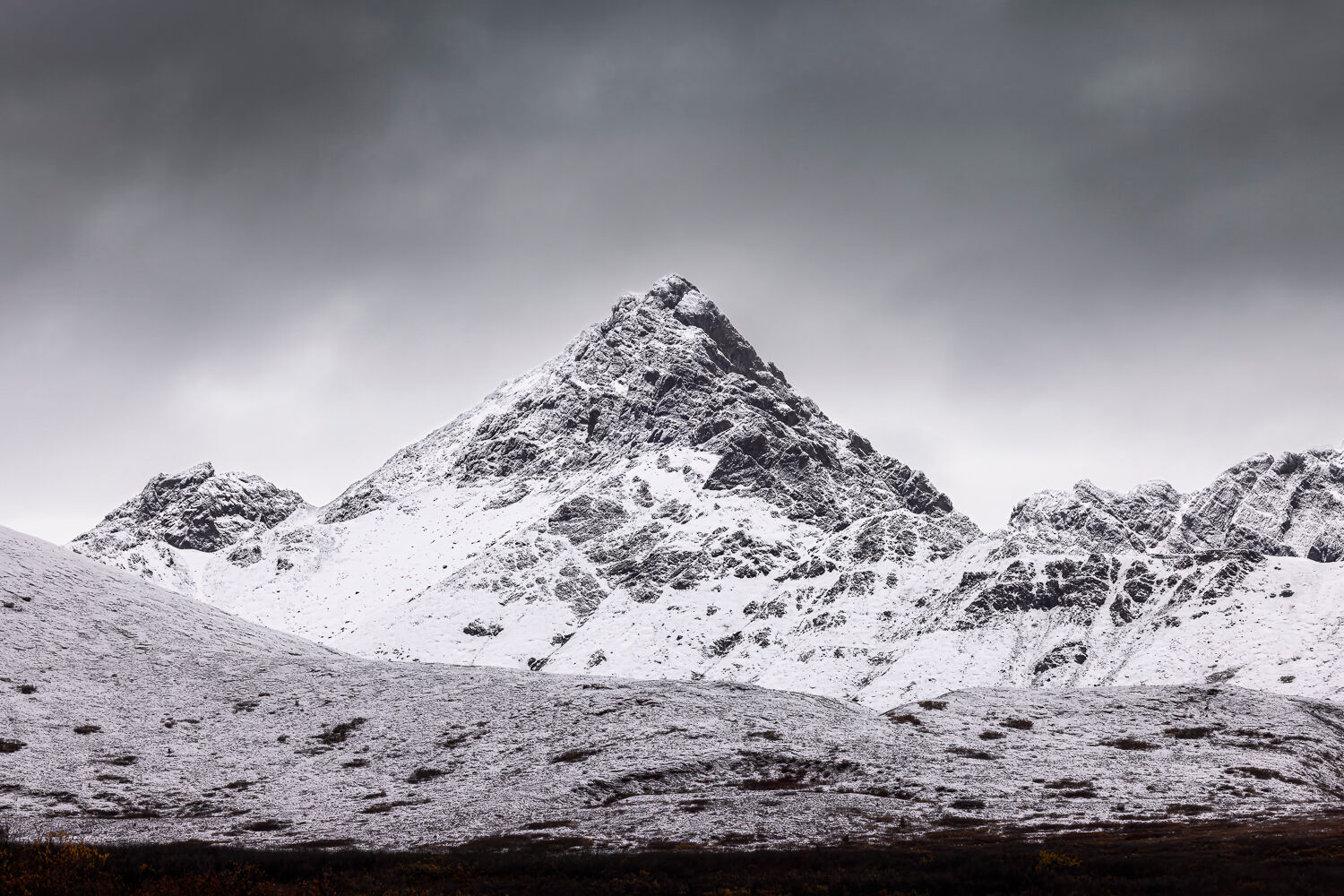 Snow-dusted mountain range in Alaska under an overcast sky, captured during a fall photography workshop. A prominent central peak rises dramatically, surrounded by rugged, snow-covered slopes and dark foreground hills. The monochromatic palette highlights the raw, serene beauty of the Alaskan wilderness.