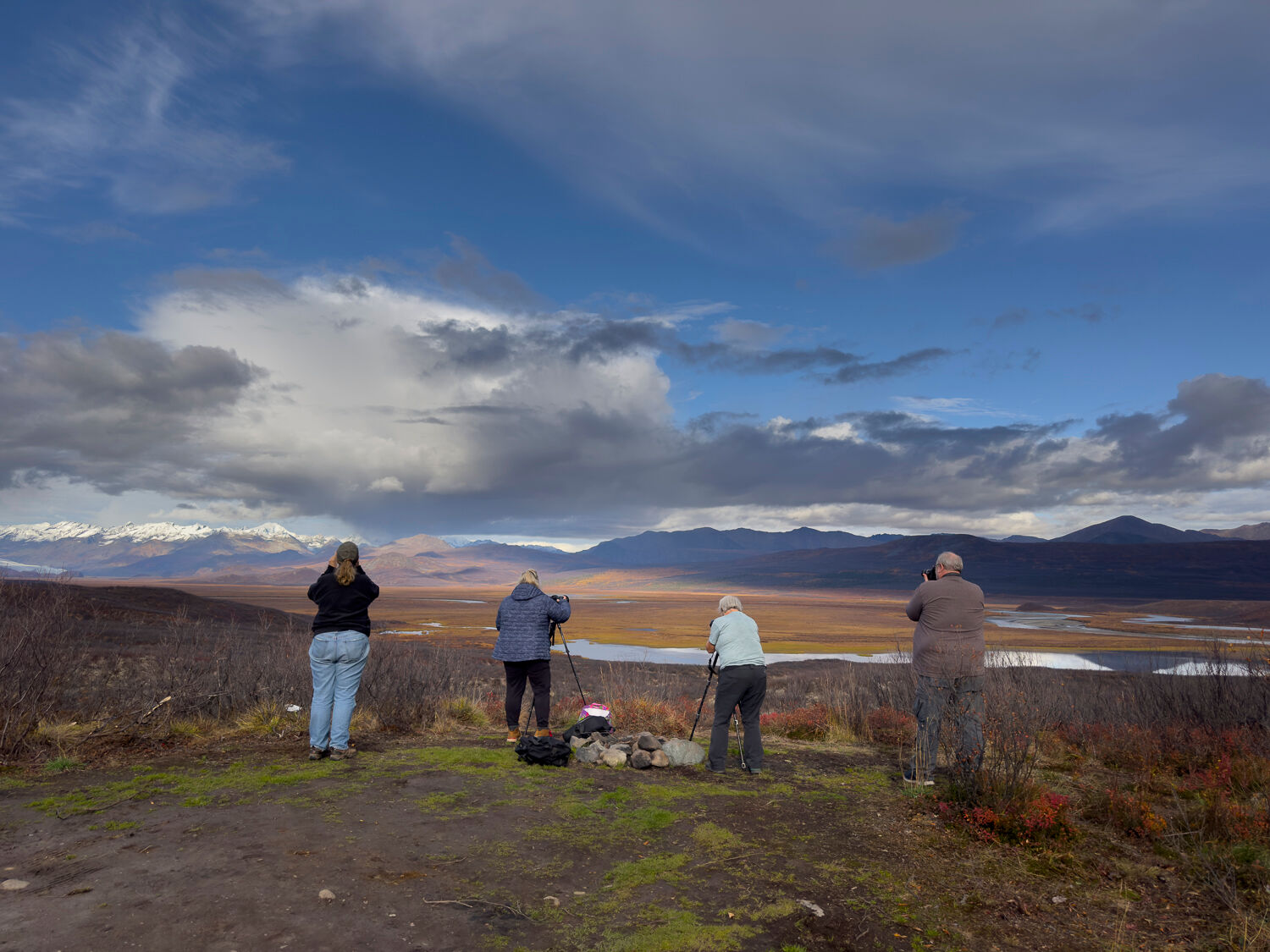 Photographers capturing Alaska's autumn wilderness during a fall photography workshop. Three individuals with tripods and cameras stand on a gravelly foreground, overlooking golden-brown plains, a reflective lake, and rugged mountains under a partly cloudy sky. The scene highlights the region's vast, scenic beauty and vibrant fall colors.