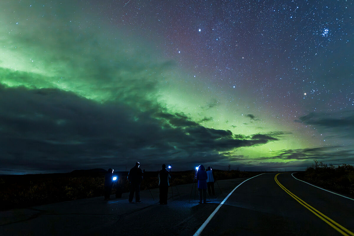 Nighttime scene of photographers capturing the Northern Lights during an Alaskan fall photography workshop. A glowing green aurora borealis stretches across the starry sky, while five silhouetted individuals with tripods stand on a curved road, illuminated by faint camera lights. The image highlights the awe-inspiring beauty of Alaska's night skies.