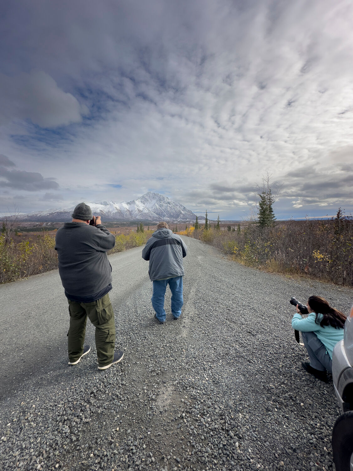 Photographers on a gravel road capturing Alaska's snow-capped mountains during a fall photography workshop. The scene features a dramatic cloudy sky, vibrant autumn foliage, and a rugged mountain range in the distance, highlighting the natural beauty of Alaska's wilderness.