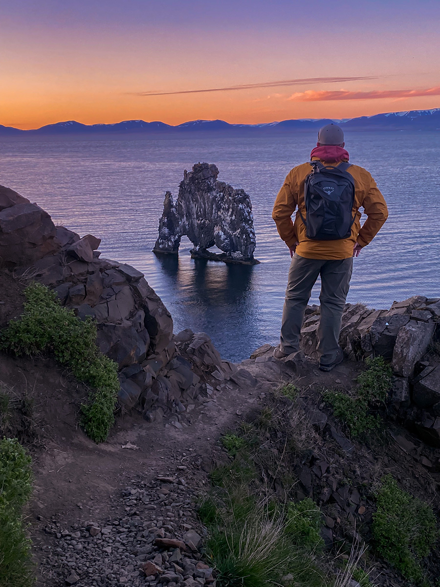A person in a yellow jacket stands on a rocky ledge, overlooking a sea stack at sunset.