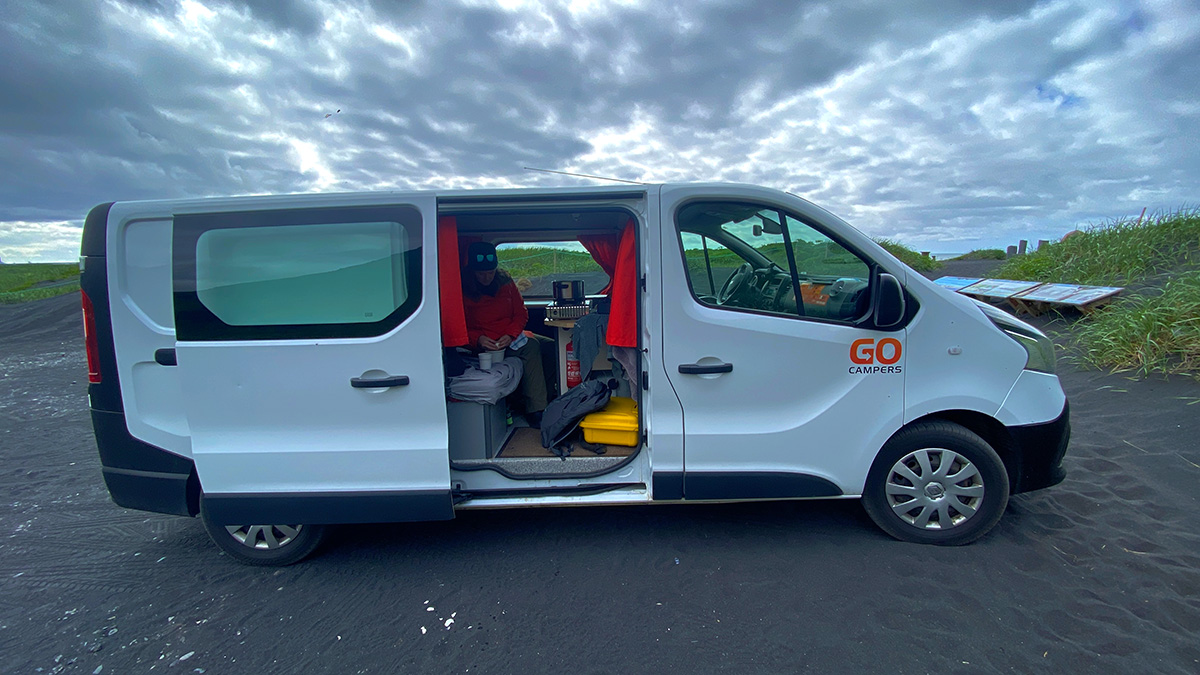 A white camper van parked on a beach with black sand, showing an open door and a person sitting inside.