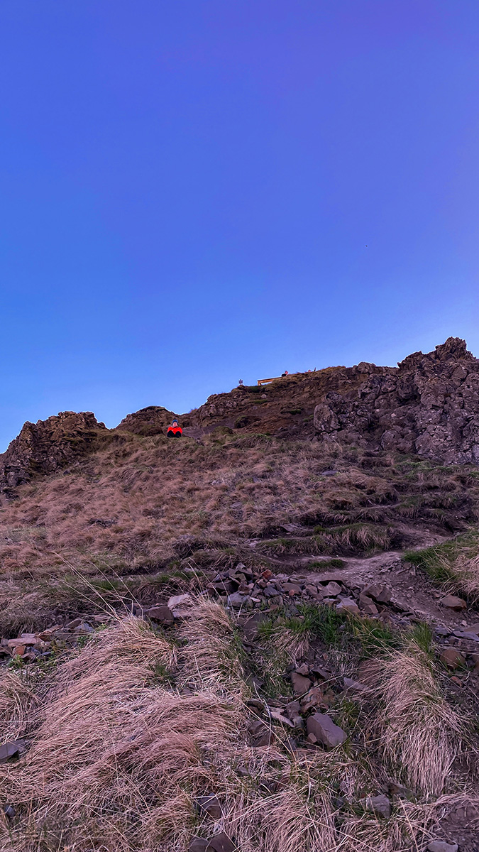 Person sitting and standing on rocky hill with grass, under a bright blue sky.