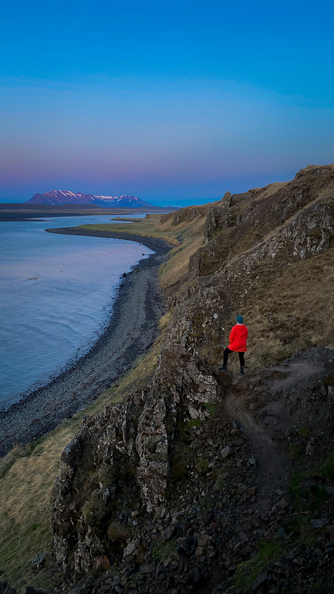 A person in a red jacket standing on rocky terrain overlooking a river and mountains.