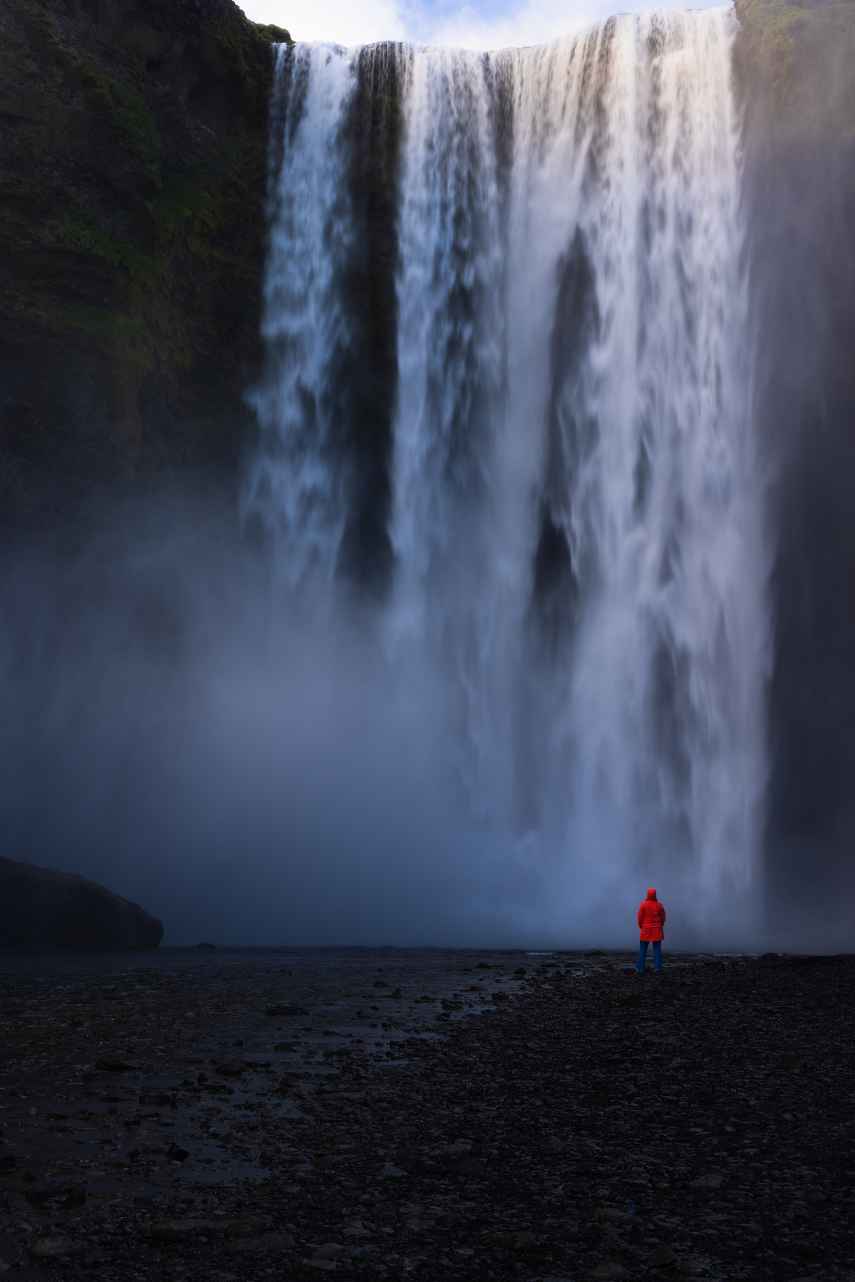 A person in a red jacket stands on a rocky shore, facing a large, cascading waterfall.