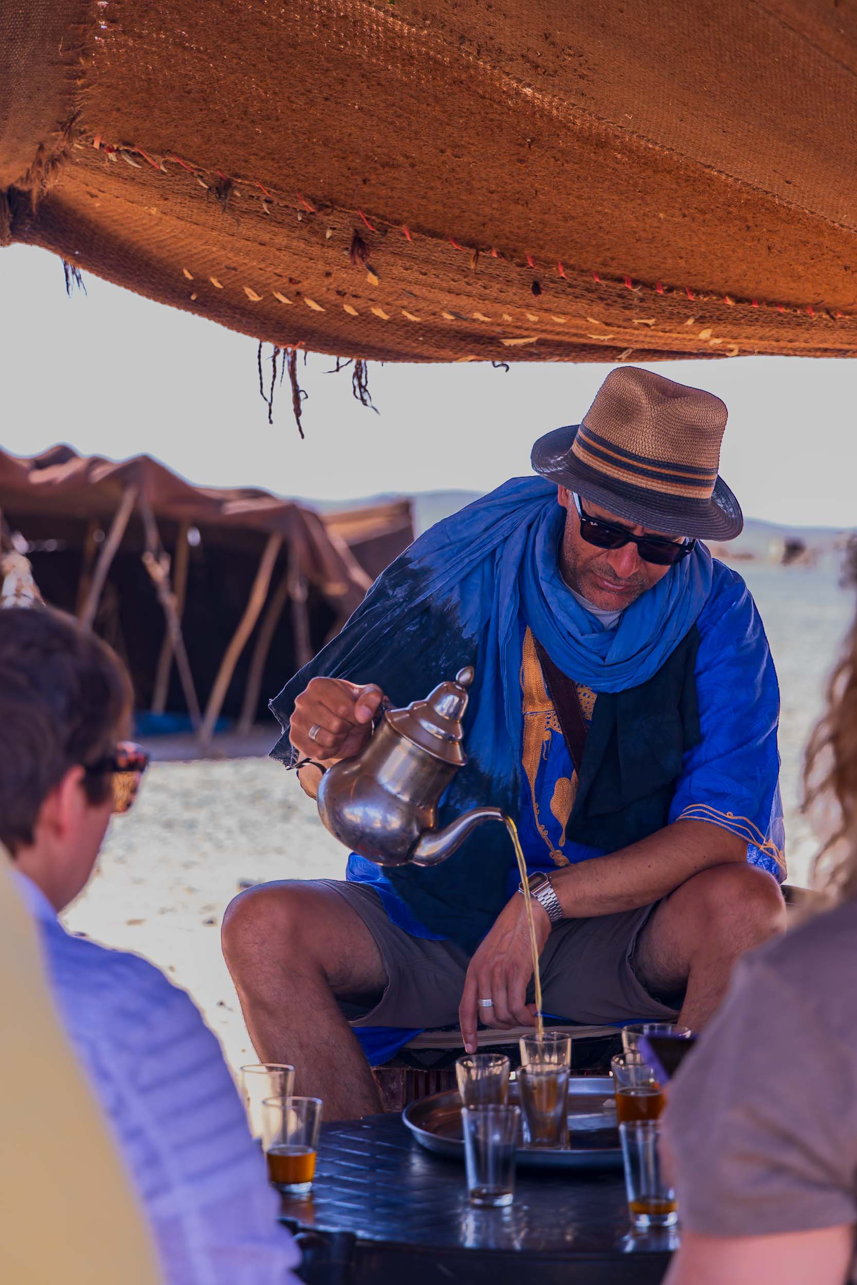 A man in a blue shirt and straw hat pours tea into glasses at a table, surrounded by people.