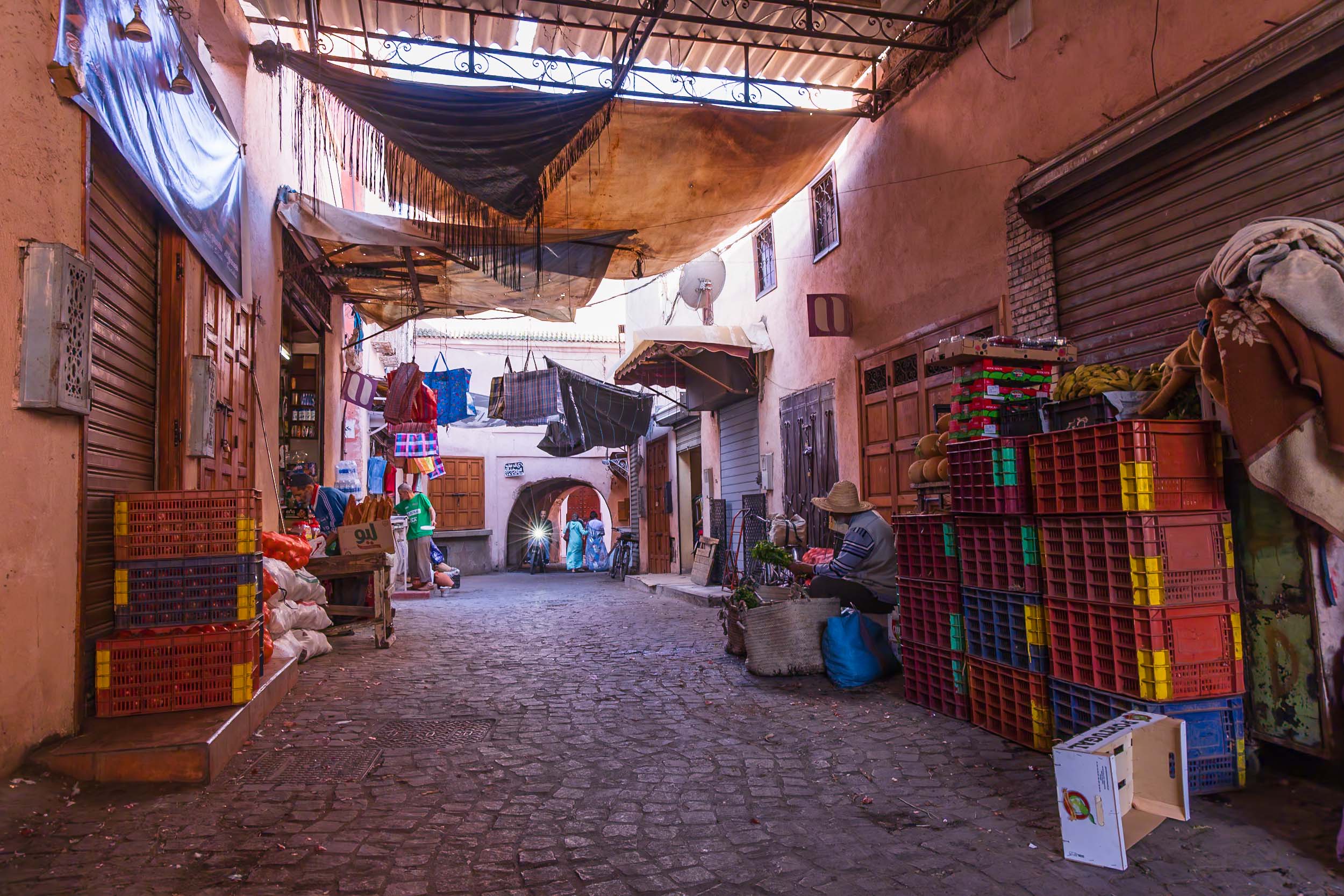 Cobblestone market street in Marrakech, lined with shaded stalls and vibrant crates as a vendor sits amidst the bustling charm.
