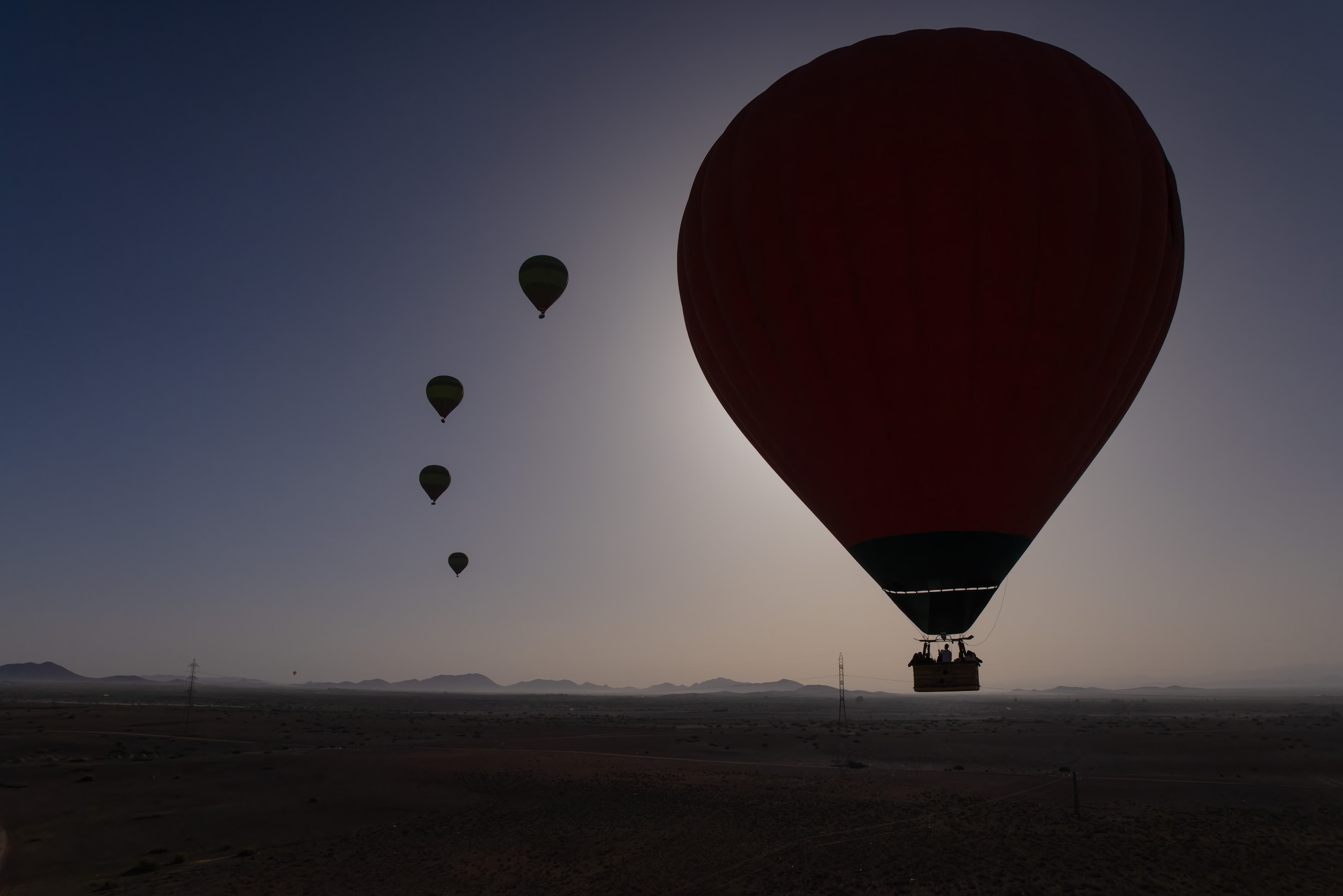 A red hot air balloon with passengers silhouetted, flying above a desert landscape with other balloons in the background.