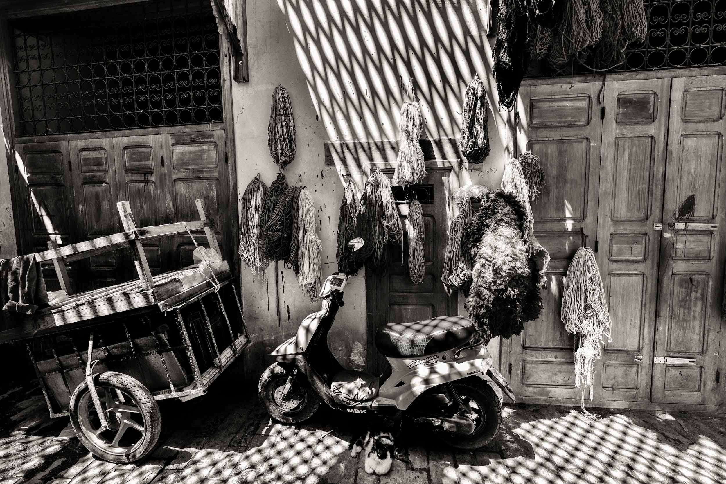 A scooter rests by a wooden cart in a shaded Marrakech alley, with ropes hanging from the wall, capturing a rustic street vibe.