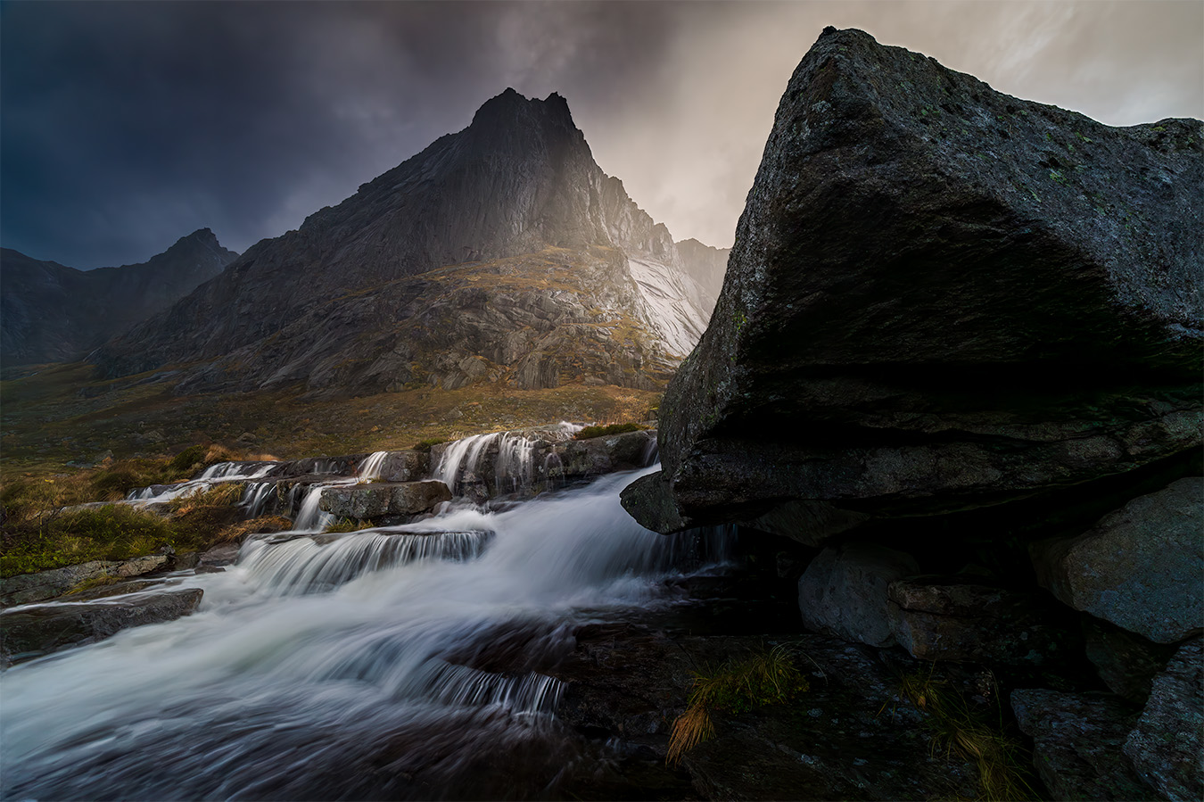 Sun glows behind the rocks under a cloudy sky lighting up the flowing water and background, with a mountain in the background.