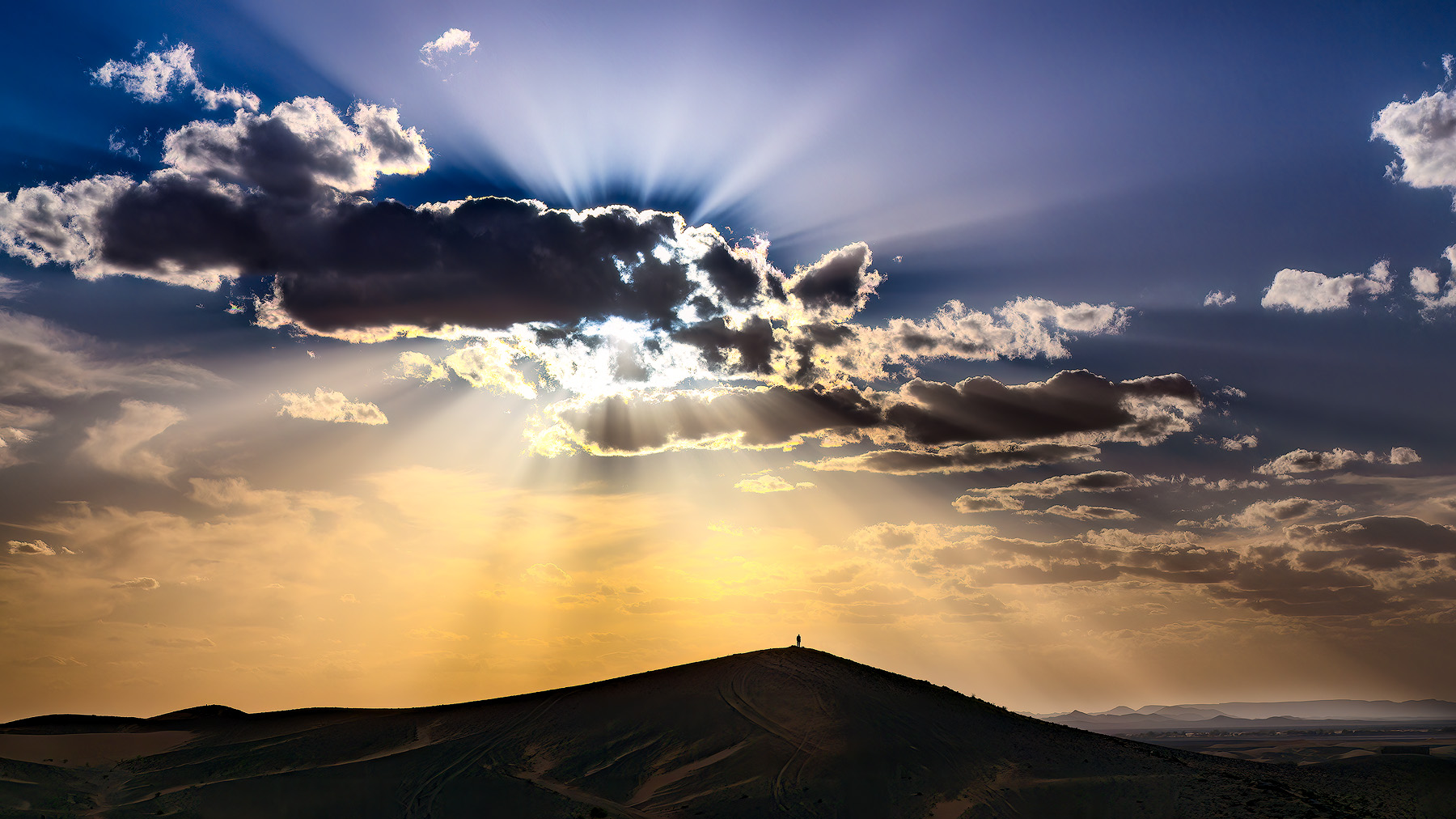 Sunbeams break through the clouds, casting light rays as a man stands silhouetted atop a hill, highlighting the vast scale of the landscape.