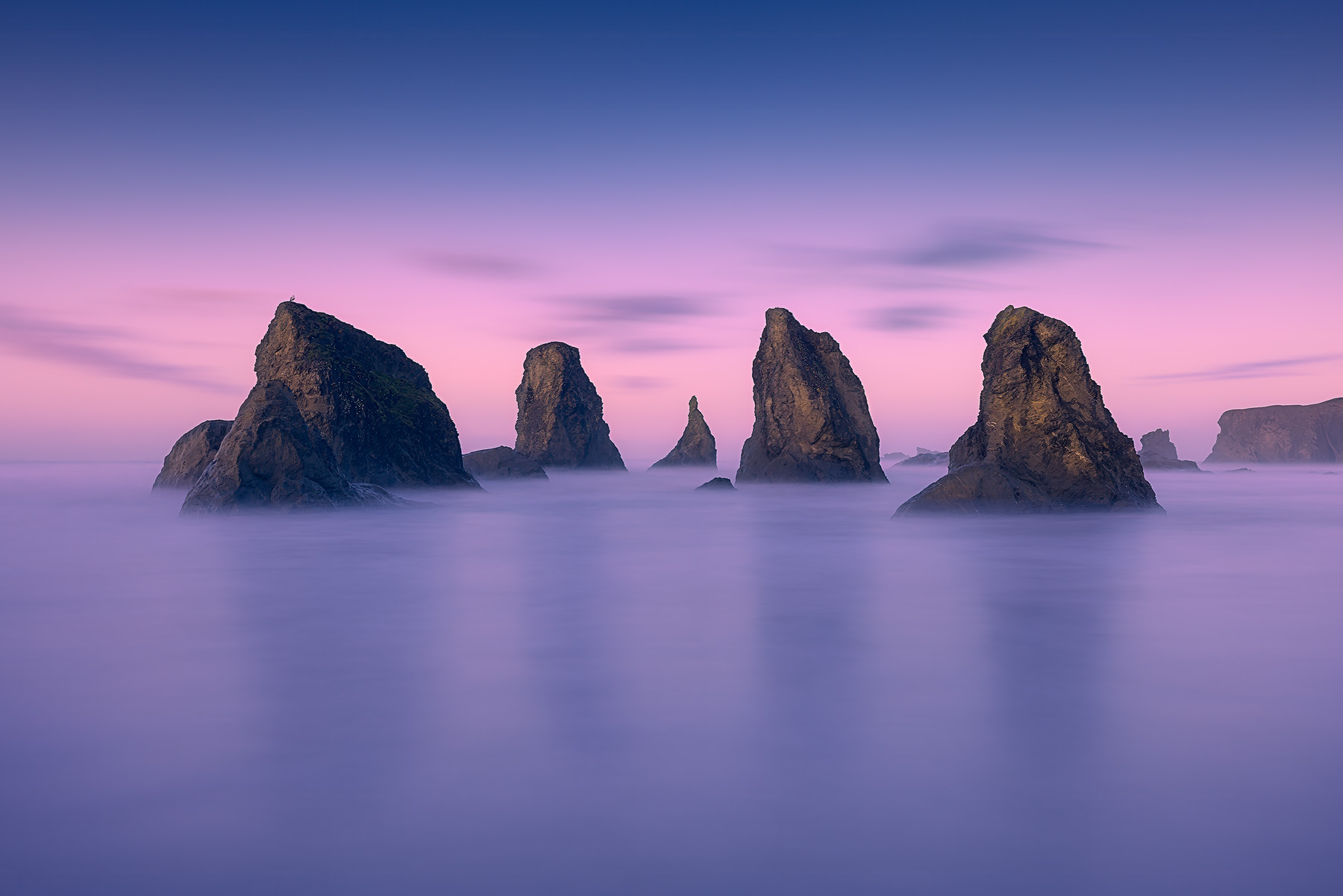 A serene long-exposure of sea stacks at sunrise on the Oregon coast, with soft surf and a peaceful, colorful sky.