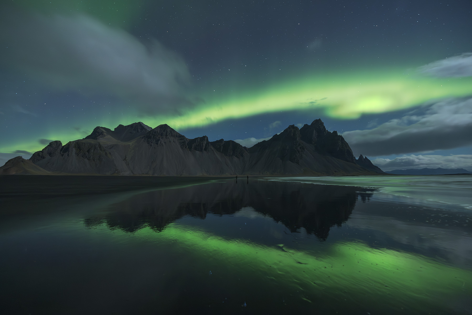 Green aurora borealis with dramatic reflection in still water at Vestrahorn mountain Iceland, with two small figures standing on black sand beach providing scale to the towering peaks and sweeping northern lights