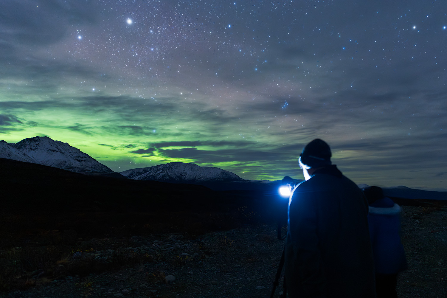 Aurora photographer silhouetted against green northern lights with headlamp checking camera settings, with snow-capped Alaska mountains and starry sky in background