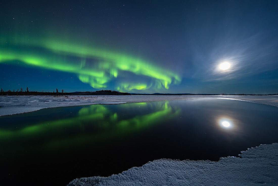 Bright green aurora borealis lights up the night sky, reflecting on a dark lake with snow-covered edges.