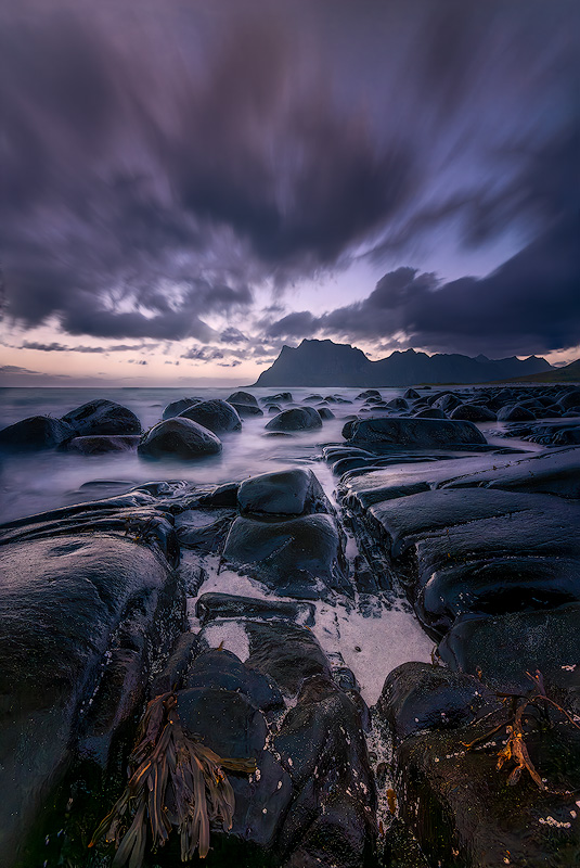 Focus-stacked seascape at Uttakleiv Beach Lofoten Norway with sharp foreground seaweed and wet rocks leading to distant mountain peaks and streaking clouds