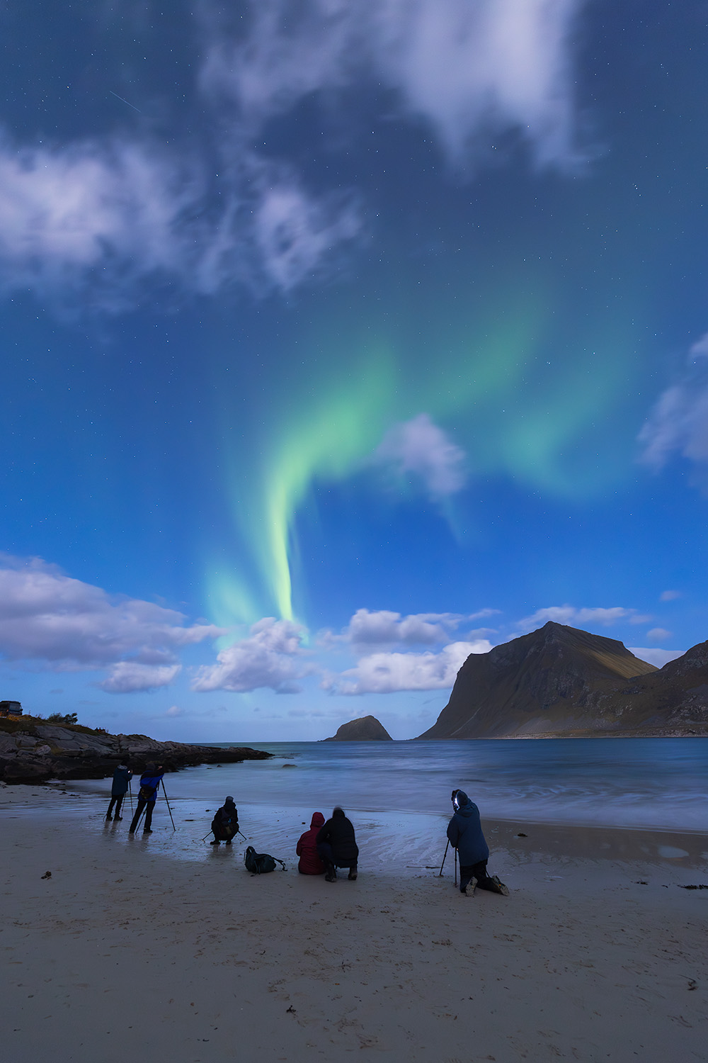 A group of photographers photograph the northern lights dancing in the sky over open water with mountains in background