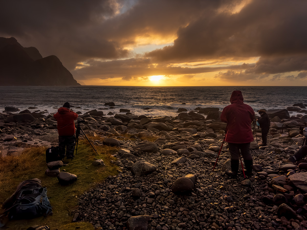 Photographers capture the landscape of Norway during sunset on a rocky beach, mountains in background
