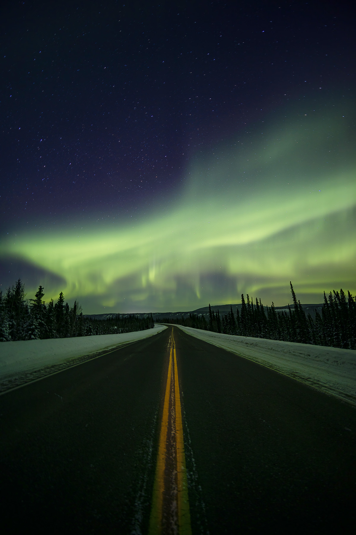 A road in Alaska stretches under a vibrant aurora borealis, surrounded by a starry sky, offering a breathtaking night view.