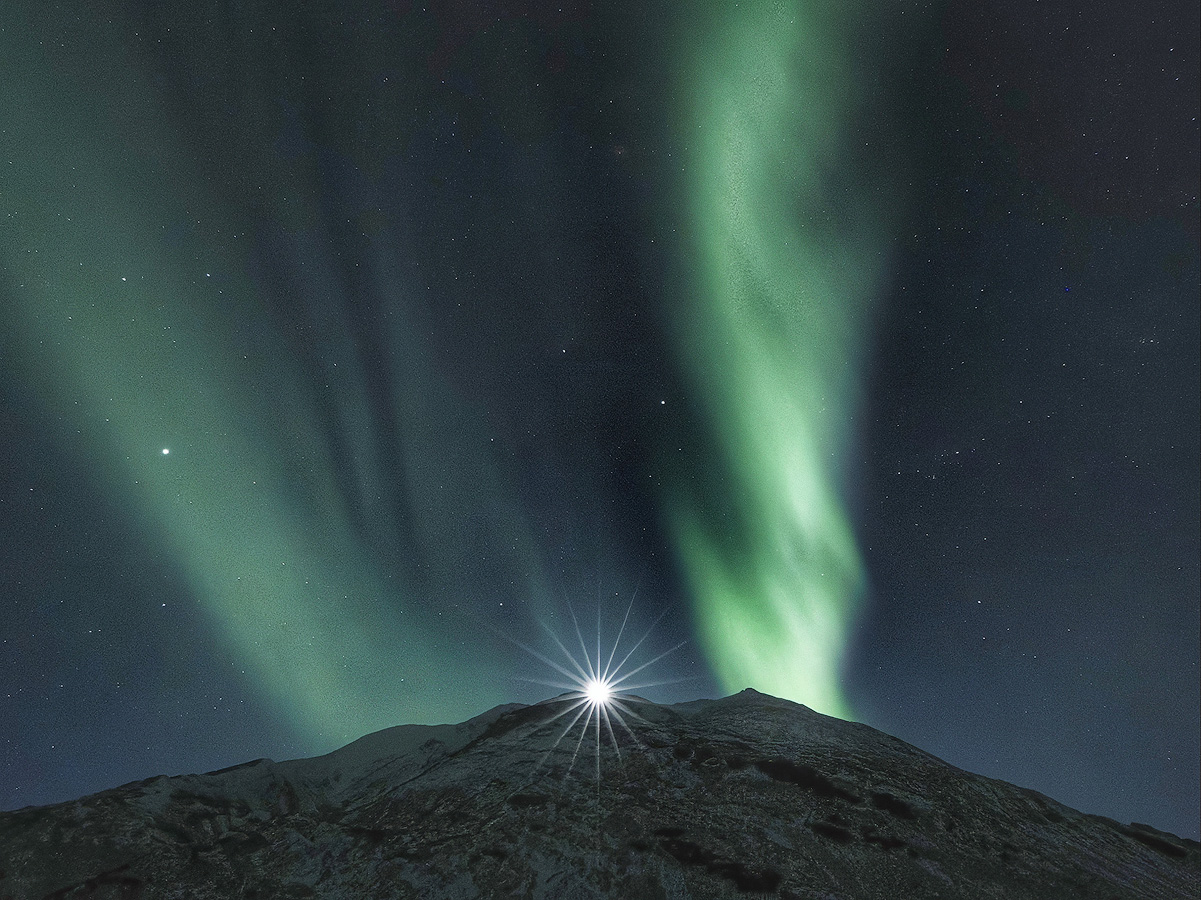 Vertical aurora curtains framing a bright moon or planet rising over a snowy mountain peak during the Spring 2026 Northern Lights Workshop in Alaska