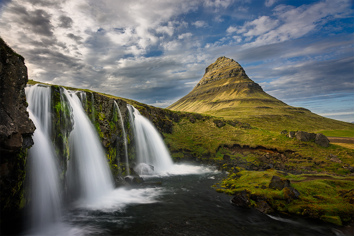 A waterfall cascading over rocky cliffs with green moss, beside a conical mountain and a cloudy sky.