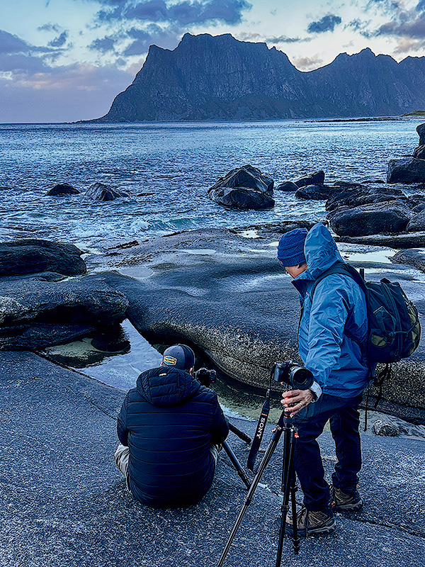 Michael Schultz crouching on rocky Lofoten coastline teaching focus stacking technique to workshop participant with tripod during blue hour scouting session
