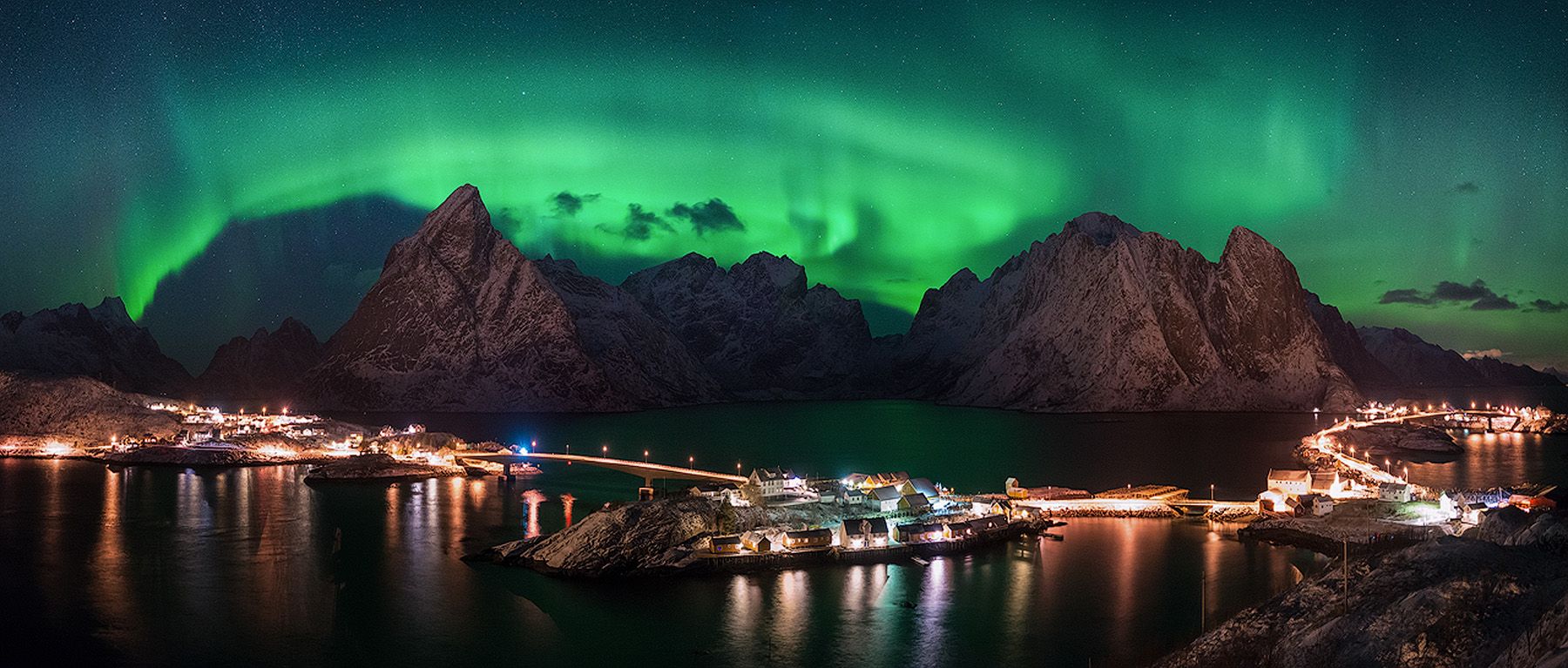 Panorama of the northern lights over a Lofoten fishing village and its lights on a winter night in Norway