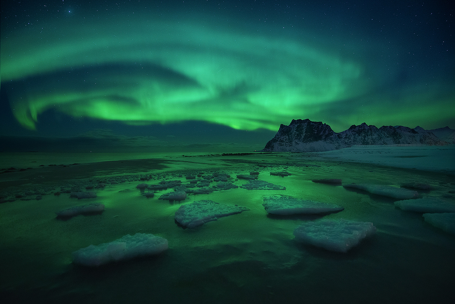 Swirling northern lights over an icy beach below snowy mountains in Lofoten, Norway in winter