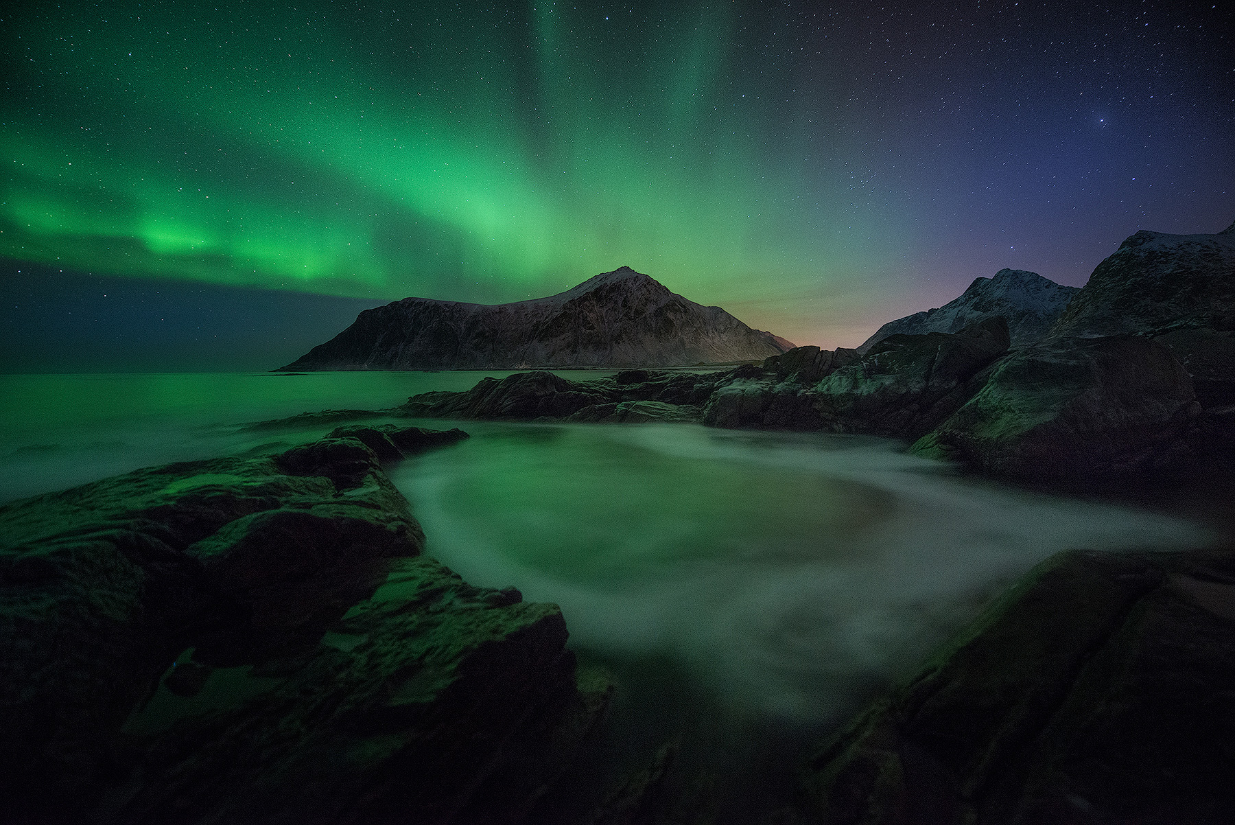 Northern lights over snow-covered mountains reflecting in a tidal pool in Lofoten, Norway on a winter aurora photography workshop