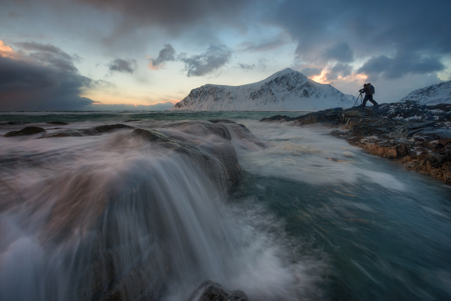 Workshop photographer on the rocks shooting a long exposure of waves on the Lofoten coast in winter, Norway
