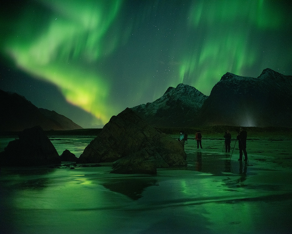 Photography workshop participants photographing aurora borealis reflected on a wet sand beach with Lofoten Islands mountains backlit in Northern Norway