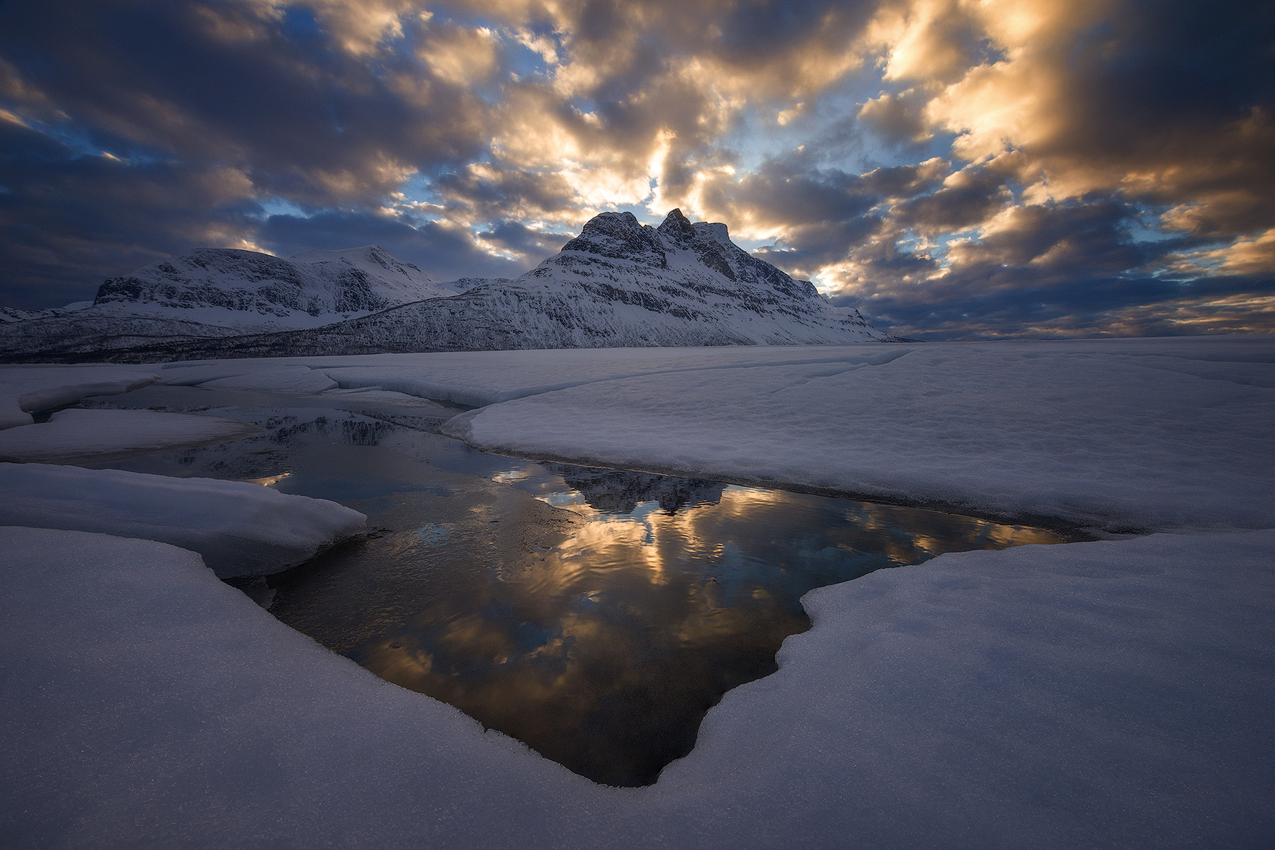 Golden storm clouds reflecting in open water on a snow-covered shore below a mountain in Lofoten, Norway in winter